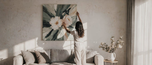 Woman of color hanging a large floral canvas print above a neutral sofa in a softly lit living room.