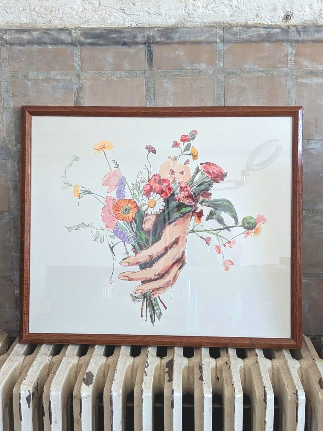 Framed artwork of a hand holding flowers on a radiator with a tiled wall background. Artist: Joëlle Diane