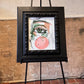 Artistic depiction of a face on a framed piece of art displayed on an easel against a textured wall. Focus on one eye. Artwork by Joëlle Diane of Monochrome Canvas.