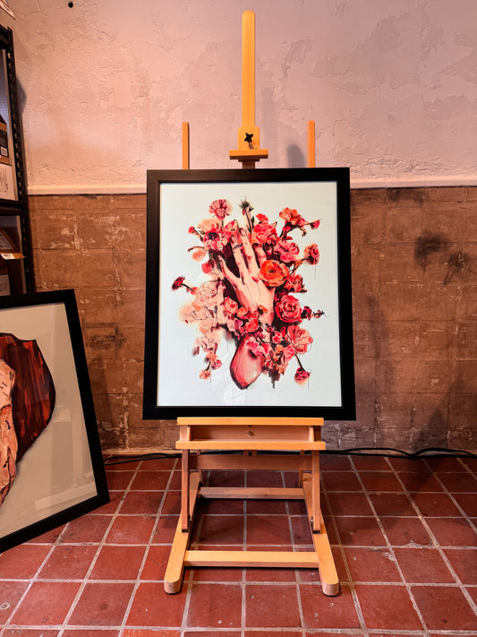 Framed artwork of a hand holding flowers on an easel against a textured wall. Joëlle Diane artwork. 