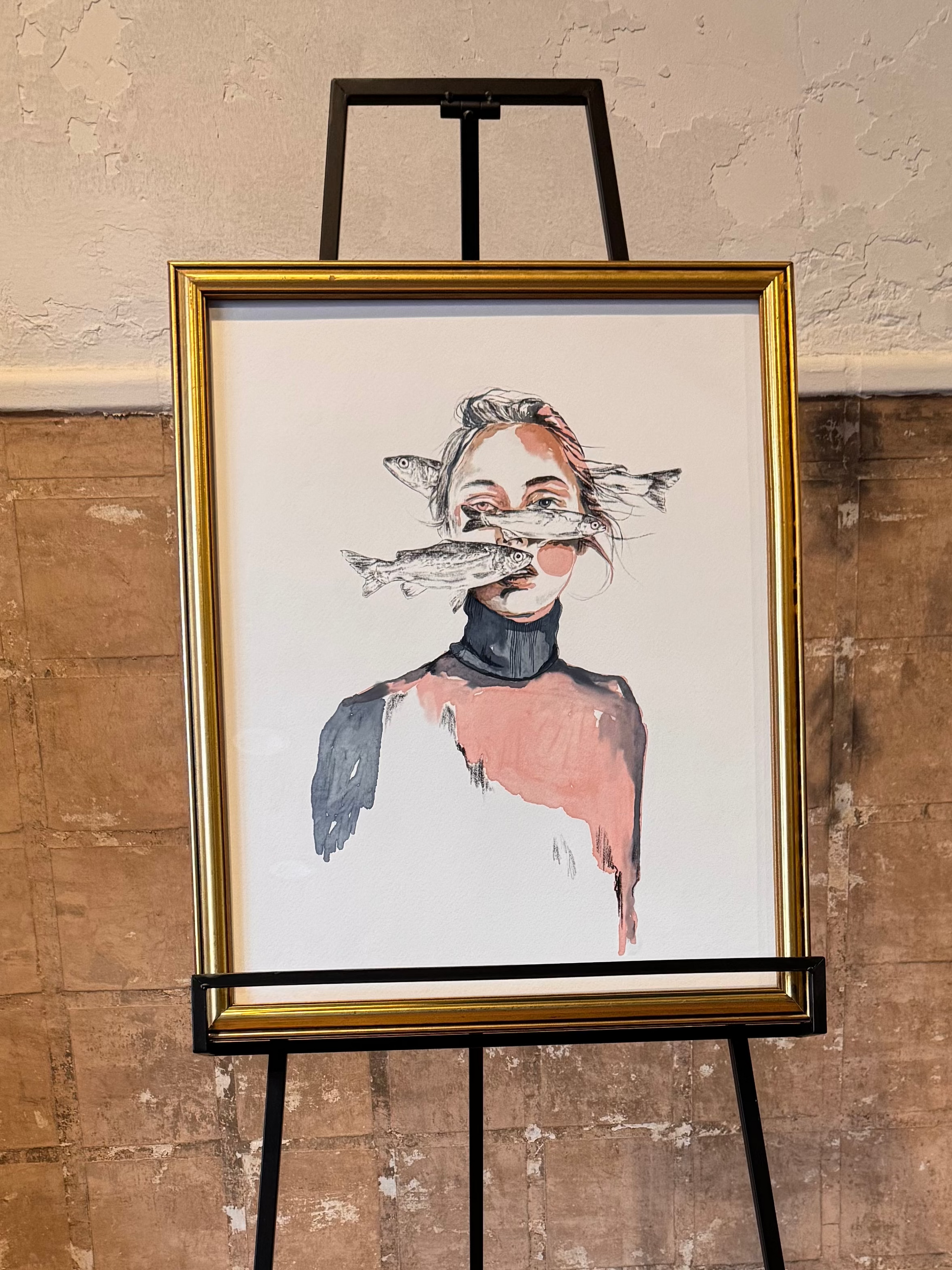 Framed artwork in a gold frame on an easel against a textured wall. Portrait of woman with fish swimming in front of her and her body disappears into the background. Artwork by Joëlle Diane of Monochrome Canvas. 