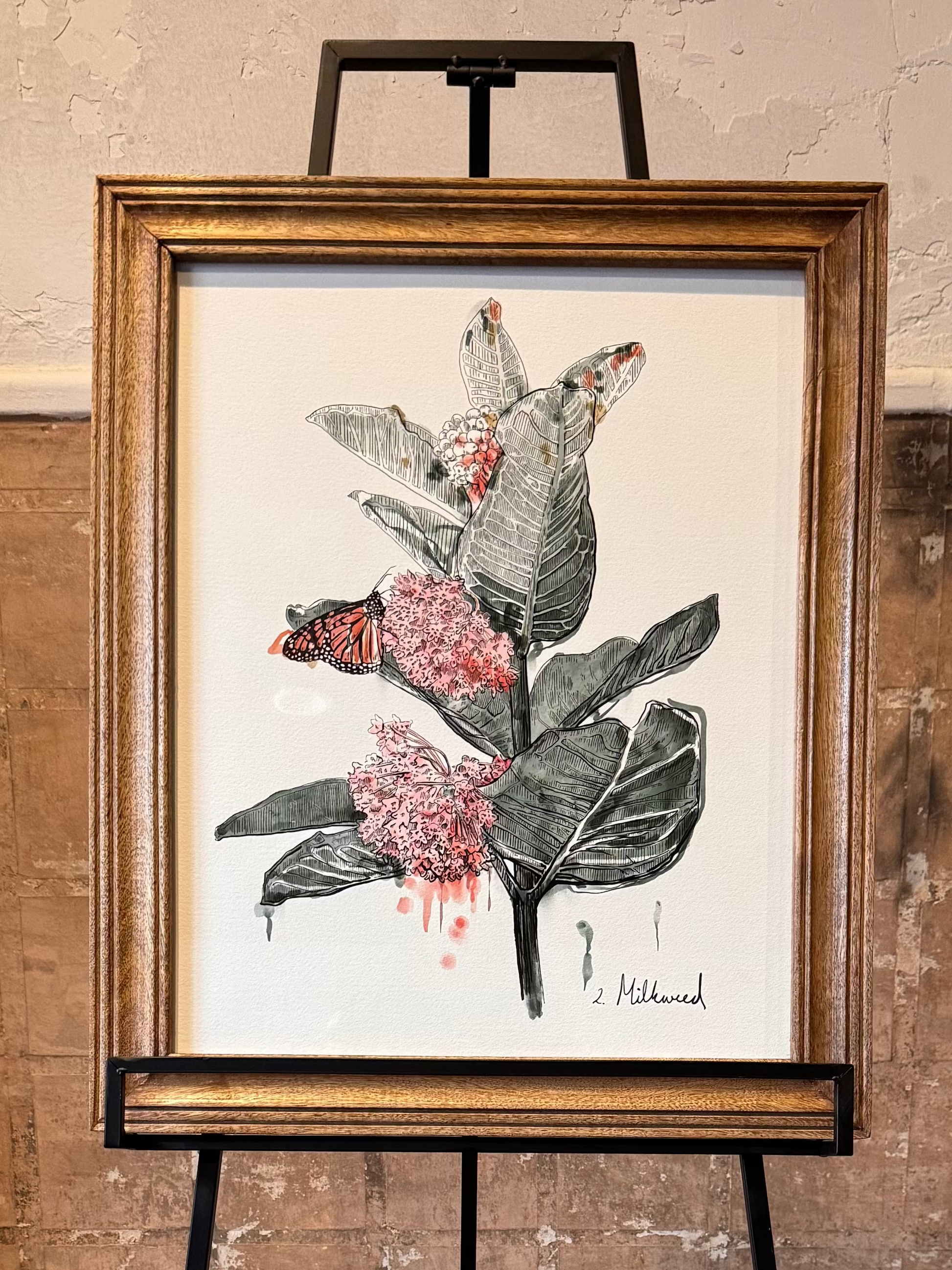 Framed artwork of monarch butterfly on milkweed plant on an easel against a textured wall. Artist: Joëlle Diane