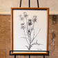Framed botanical illustration on an easel against a textured wall. Thistle artwork. Artist: Joëlle Diane