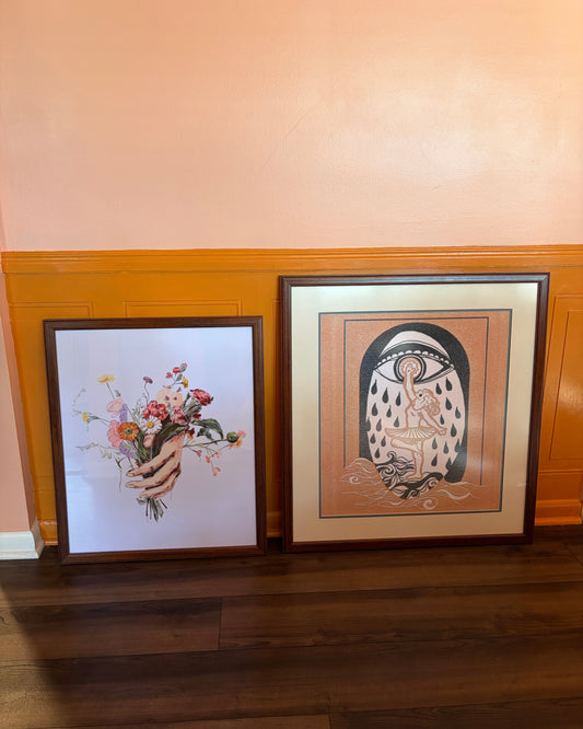 Two framed art prints leaning against a peach-colored wall with wood wainscoting and dark hardwood floors. The left print shows artwork "Ethogram:Override", artwork "Heart By Water"