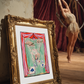 A framed art print depicting a circus performer hanging from ropes, styled in an old gold frame leaning against a wall in a rustic, dimly lit studio with a blurred circus performer in the background.