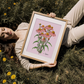 Woman lying in a field holding a framed illustration of coneflowers on a pink background